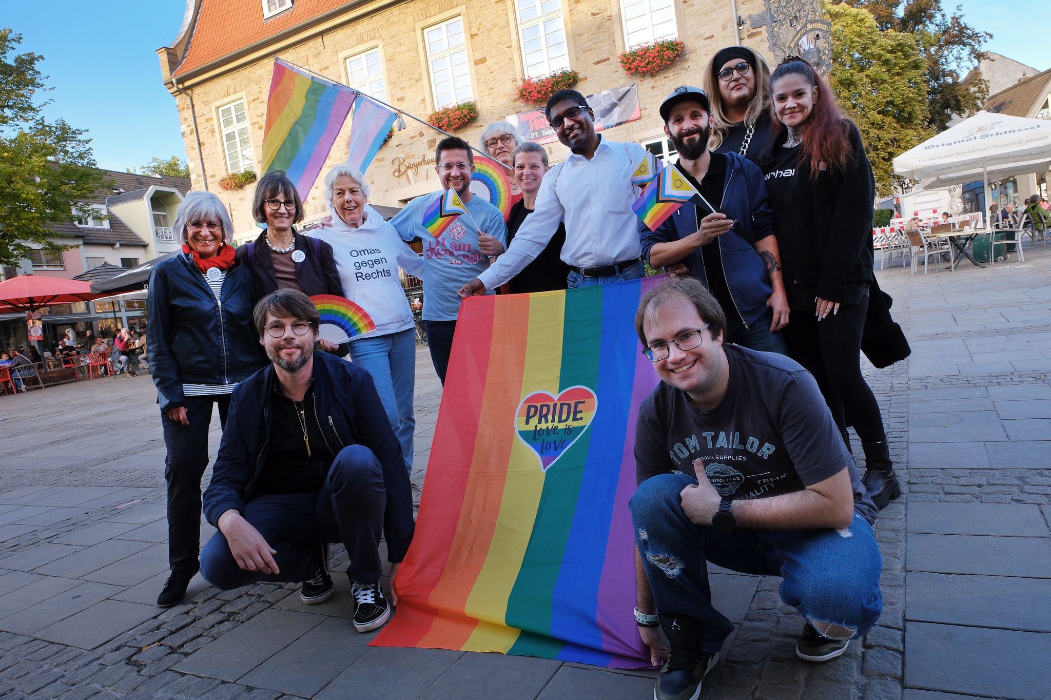 Eine fröhliche Gruppe von Menschen steht zusammen auf dem Marktplatz von Ratingen. Sie halten Regenbogenfahnen und Banner mit der Aufschrift ‚Pride – Love is Love‘ hoch. Im Hintergrund sind ein altes Gebäude (das Bürgerhaus Ratingen) mit Blumenkästen und Straßencafés zu sehen. Die Gruppe besteht aus jüngeren und älteren Personen, einige tragen T-Shirts mit politischen Botschaften wie ‚Omas gegen Rechts‘. Alle lächeln und zeigen ihre Unterstützung für Vielfalt und Gleichberechtigung.